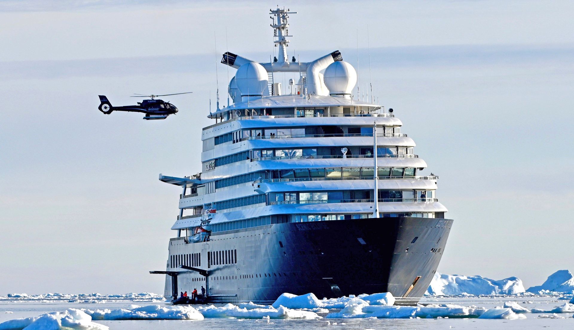 Scenic Eclipse, dark hull and white upper decks, among sea ice in Antarctica with one of its helicopters flying alongside