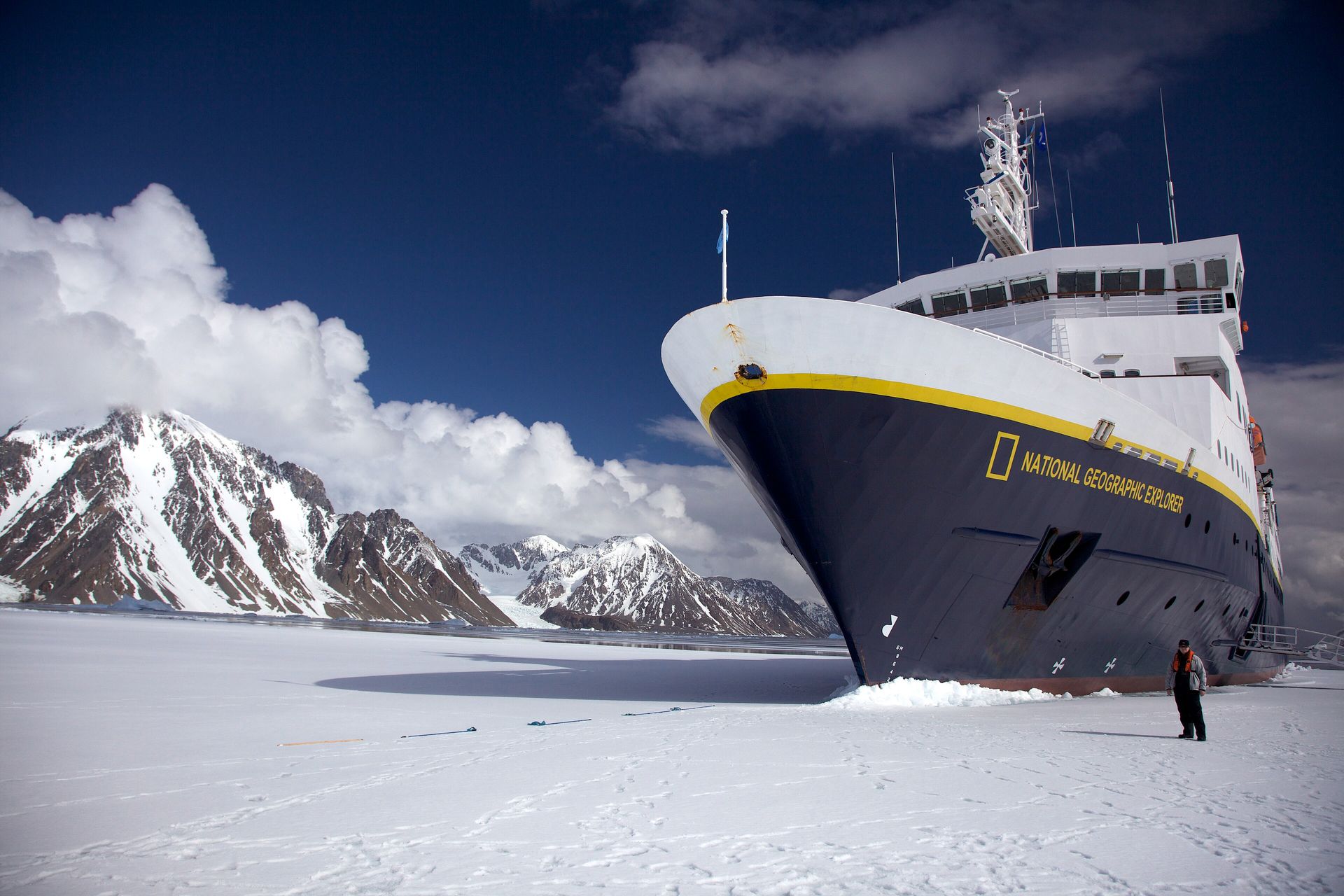National Geographic Explorer, navy hull with a yellow stripe, parked in fast ice near the Antarctic coast with a person standing on the ice and mountains behind