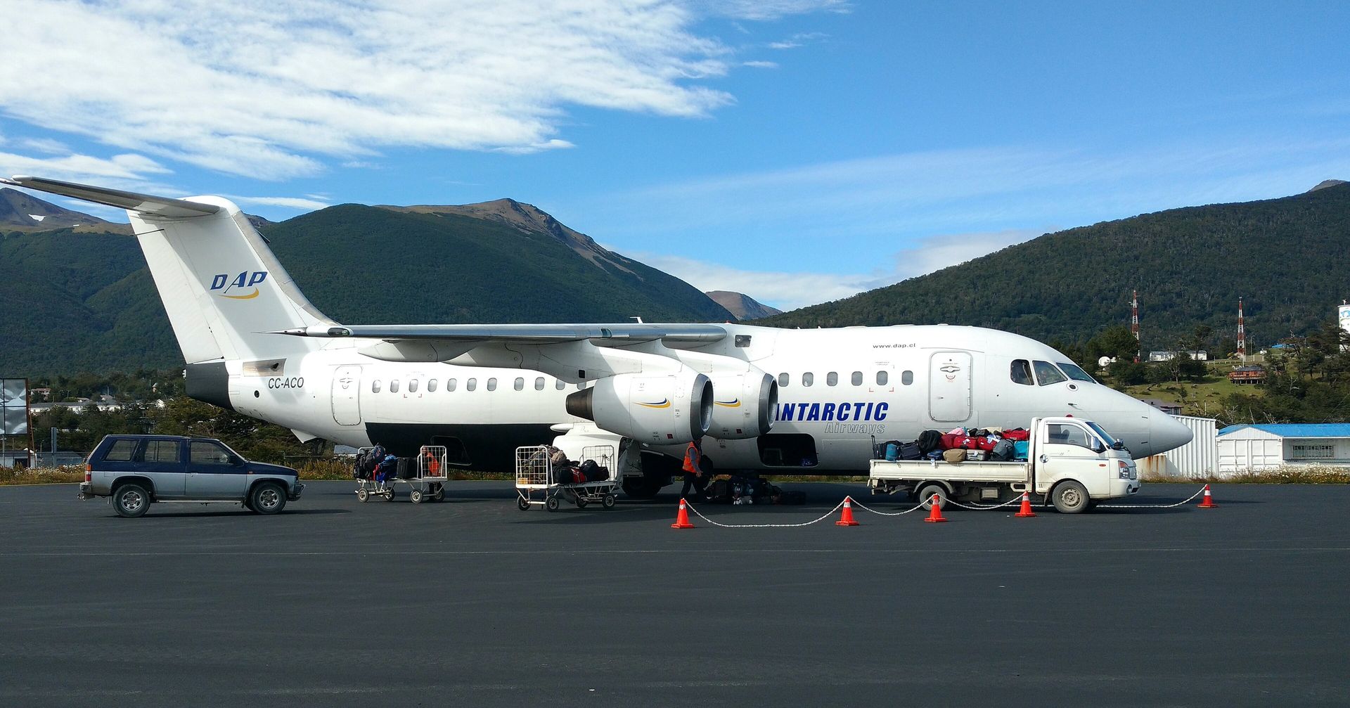 A white BAe 146 jet of Aerovías DAP in Antarctic Airways livery parked at Puerto Williams airfield, Chile, with luggage carts and a baggage truck beside it and green mountains behind