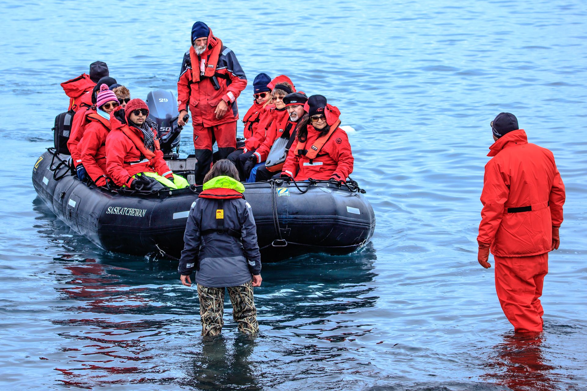 Passengers in red expedition parkas, hats and sunglasses arriving by Zodiac at Turret Point on King George Island, with guides in waterproofs standing in the shallows