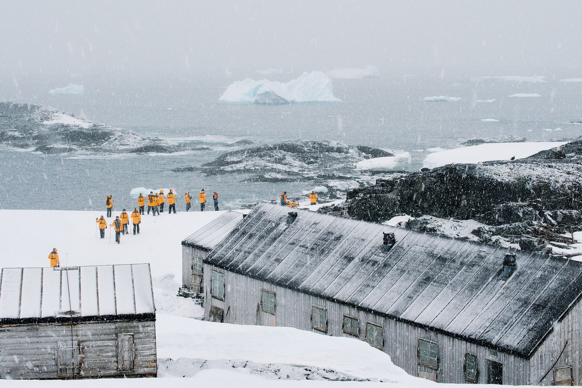 Visitors in yellow parkas walk past the wooden huts of abandoned British Base W on Detaille Island, south of the Antarctic Circle, as snow falls over icebergs
