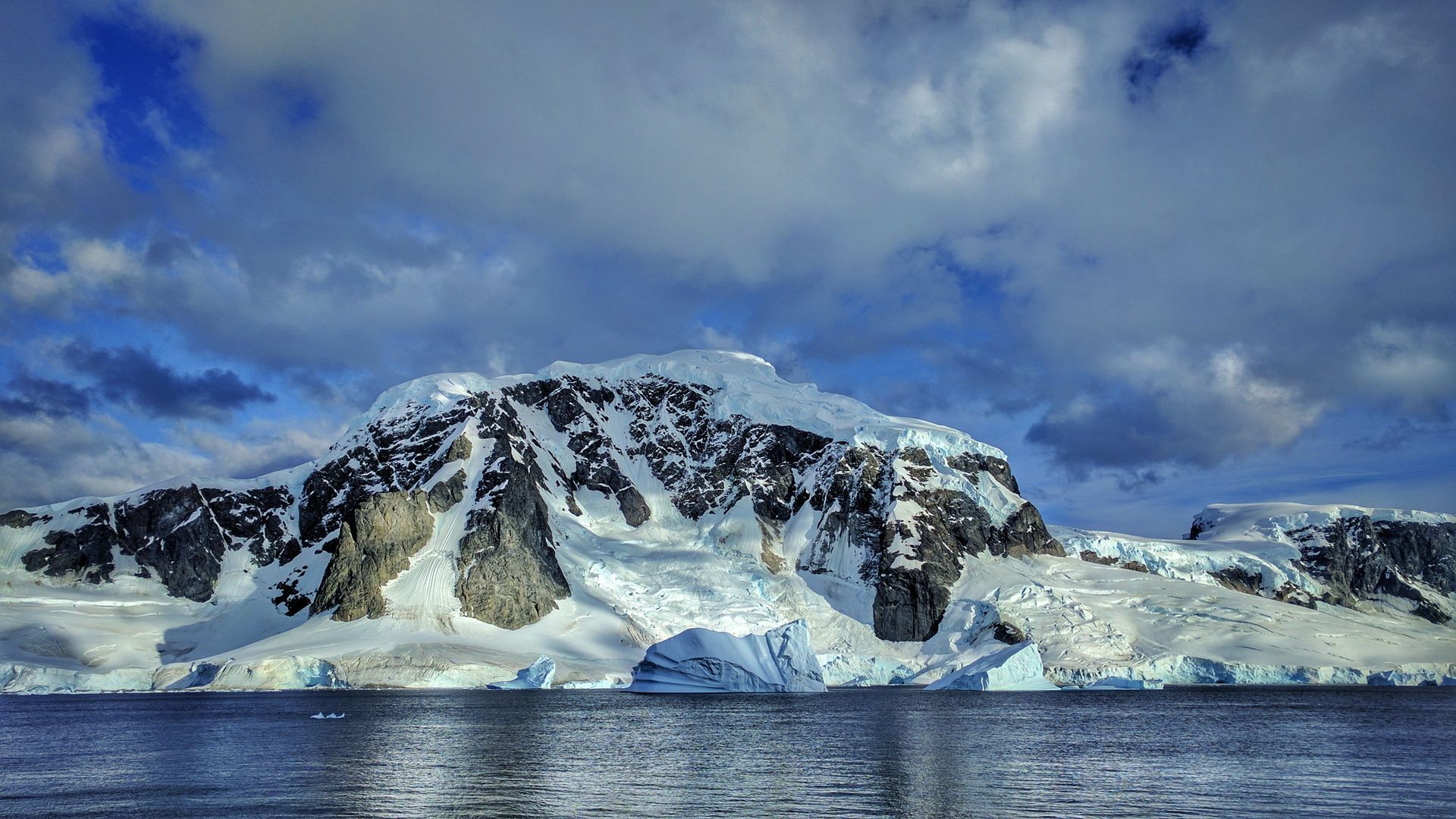 Snow and glaciers cover the dark rock of Rongé Island above an iceberg and calm water off the Antarctic Peninsula