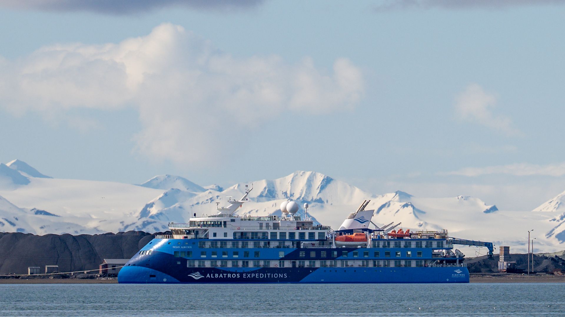 The X-Bow expedition ship Ocean Albatros, its blue hull still painted with the Albatros Expeditions name, at anchor below snow-covered mountains in June 2025
