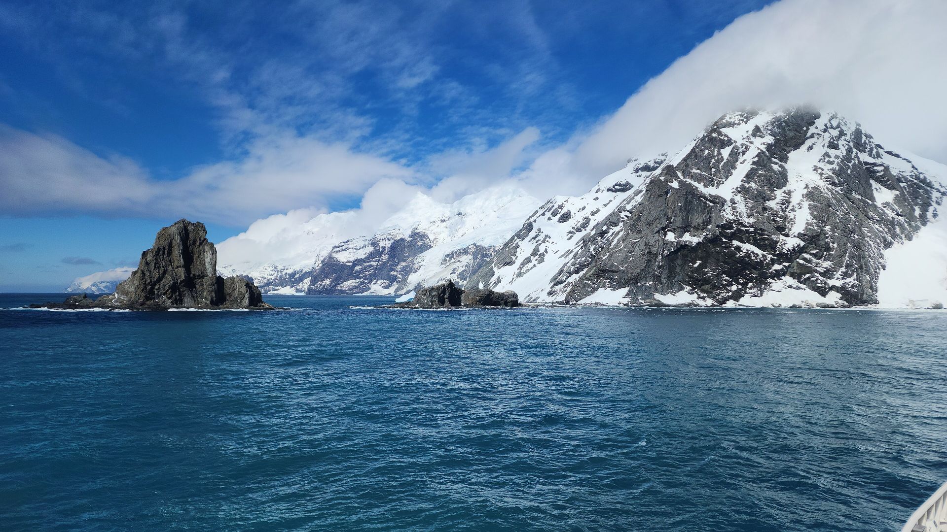 Dark rock stacks and snow-covered cliffs at Point Wild on Elephant Island, seen from a ship under a blue sky with cloud on the summit