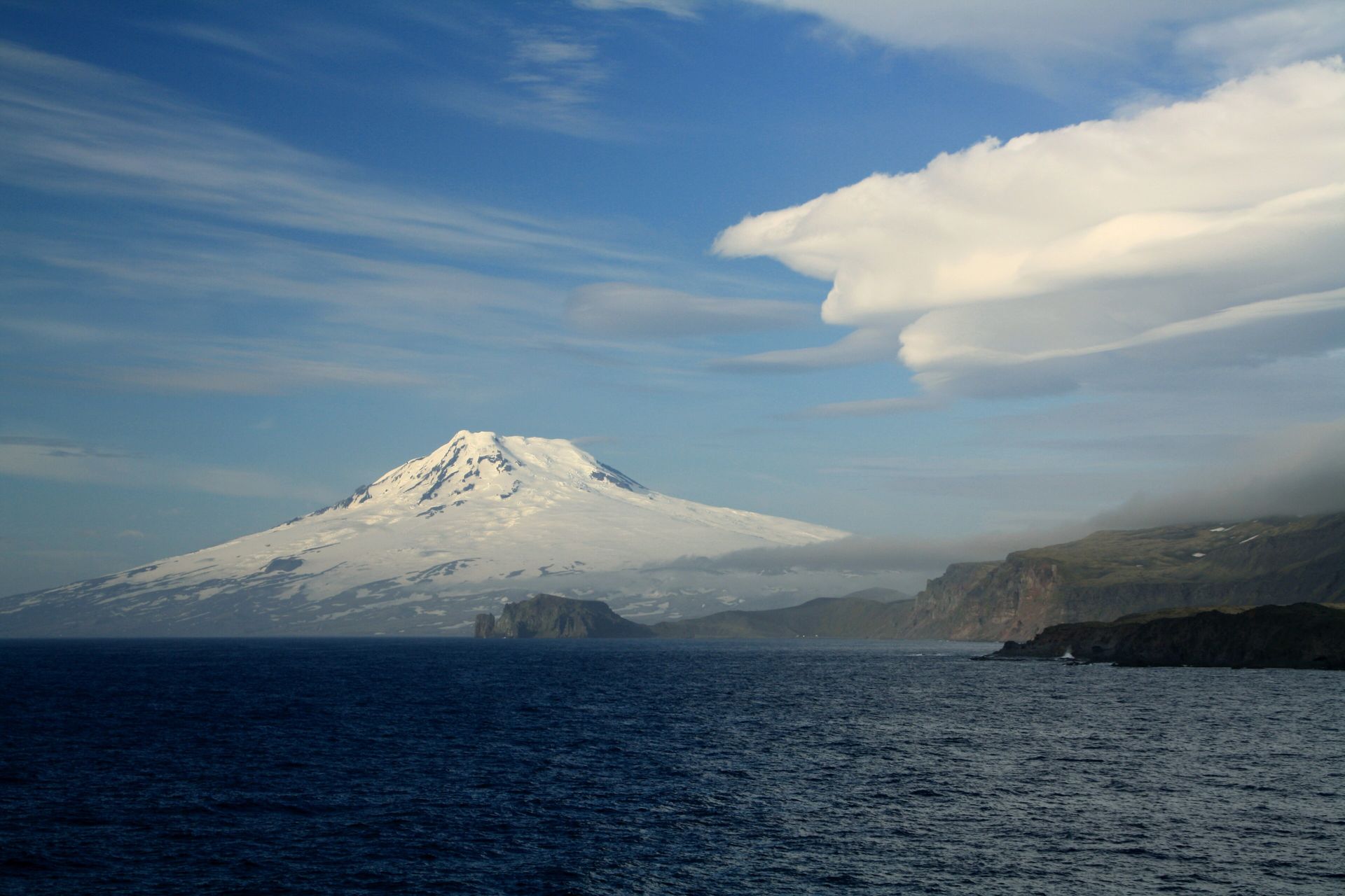 Snow-covered Beerenberg volcano rising above the dark sea and coastal cliffs of Jan Mayen under a blue sky
