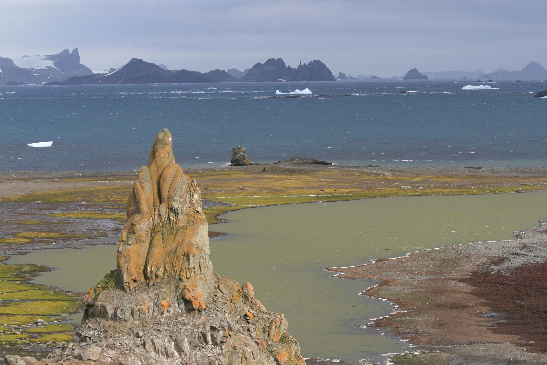 A lichen-orange rock spire above mossy green flats and a meltwater lagoon on the Aitcho Islands, with dark island peaks across the water