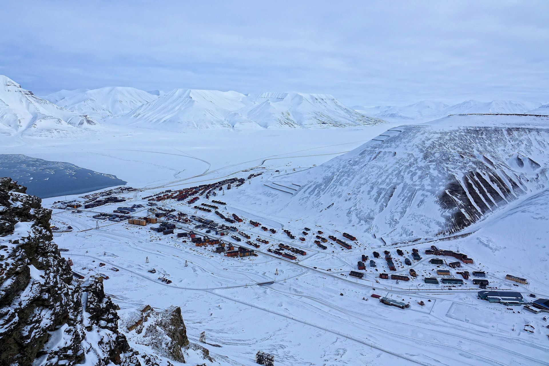 Snow-covered Longyearbyen seen from above, its colored houses spread below Sukkertoppen mountain, with the frozen Adventdalen valley stretching away behind the town