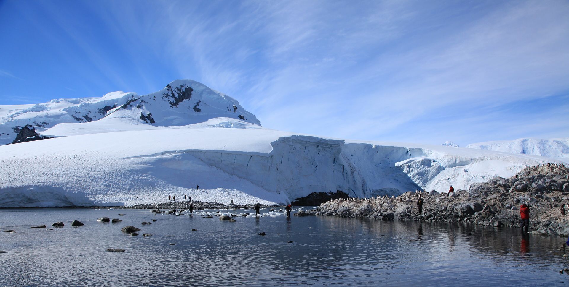 Visitors in parkas walking the shore at Waterboat Point on the Antarctic Peninsula, below an ice cliff and a snowy peak, with penguins on the rocks