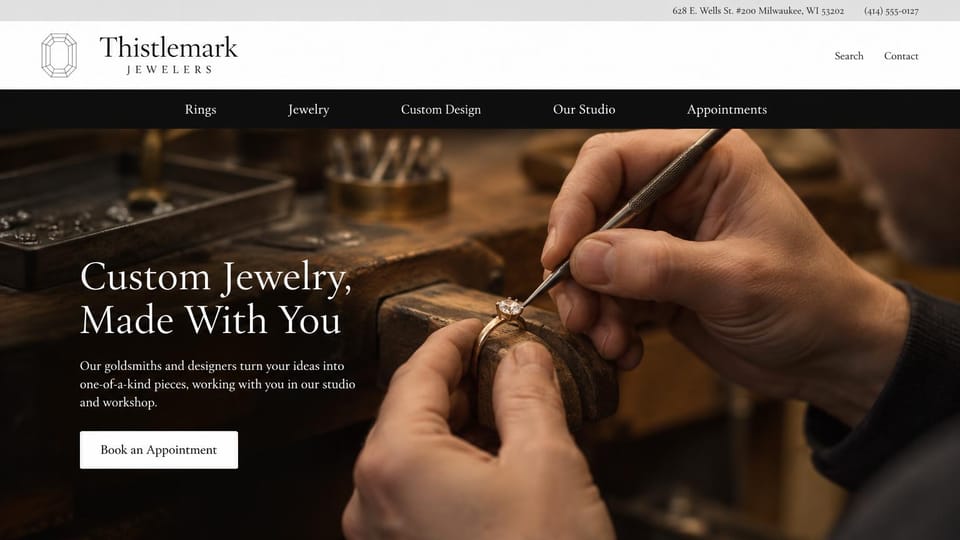 Jewelry website design, custom jewelry studio with the goldsmith's bench: gray top bar with an address and phone, white header with a serif wordmark and an emerald-cut gem outline, black menu bar, hands setting a diamond in a gold ring at a wooden bench, white serif headline, white appointment button