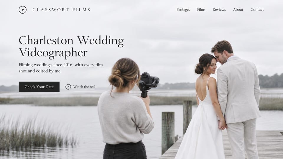 Videographer website design, solo wedding filmmaker on an overcast marsh dock: soft gray sky over a salt marsh, a camerawoman filming a couple in white and linen on a wooden dock, large black serif headline on the left, black rectangular button beside a play-icon reel link, widely spaced serif logo and menu