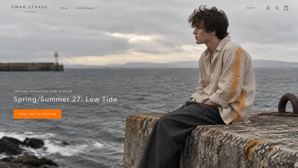 Fashion website design, coastal menswear by a lighthouse: overcast harbor, a young man in a striped linen shirt on a stone wall with a lighthouse behind, a white headline and an orange button