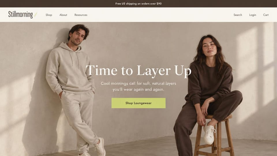 Fashion website design, loungewear for two in a sunlit studio: brown top bar, a man in an oatmeal hoodie and joggers against a plaster wall and a woman in a brown sweatsuit on a stool, a white serif headline and a lime button