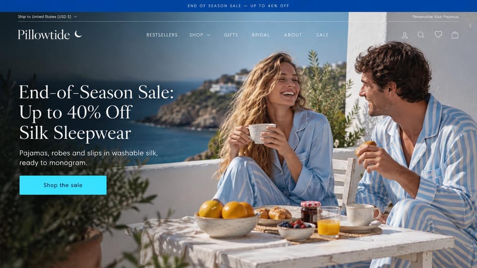 Fashion website design, silk sleepwear at a seaside breakfast: royal blue top bar, a laughing couple in pale blue silk and striped pajamas at breakfast on a white terrace above the sea, a white serif headline and a cyan button
