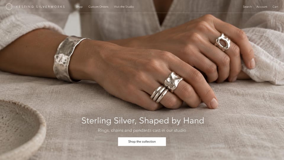 Jewelry website design, handmade silver with studio visits: taupe linen photo of hands wearing hammered silver rings and a cuff, a centered white headline and a white button