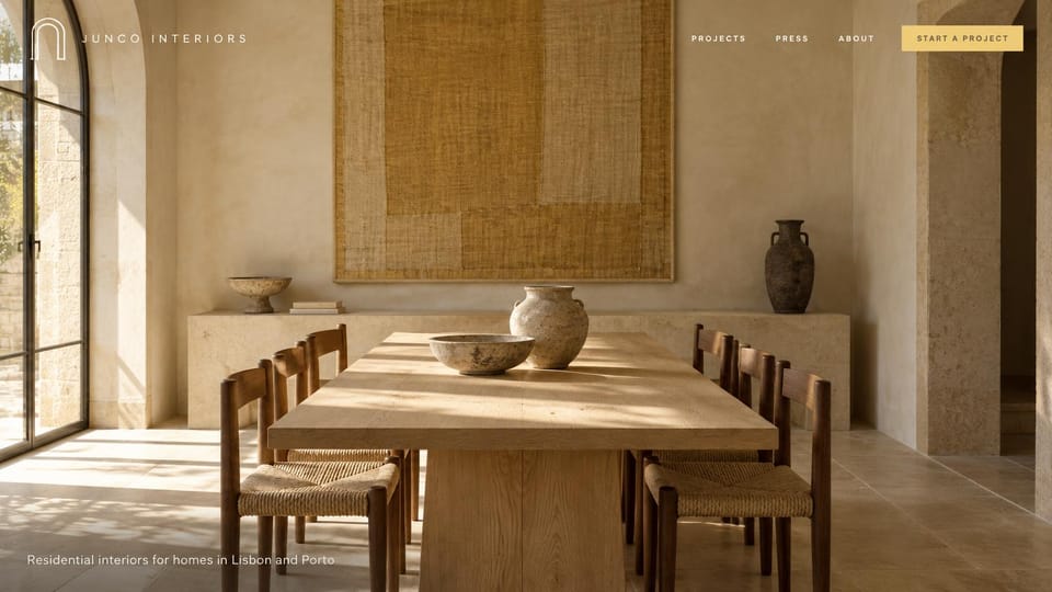 Interior design website design, warm Mediterranean homes, photo first: full-screen sunlit dining room with stone walls, an arched steel window, rush seat chairs and a woven wall hanging, spaced white capitals and a gold button
