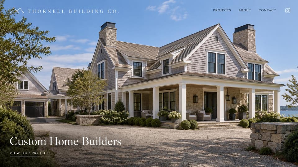 Construction website design, shingle-style custom homes by the water: a gray shingled house with stone chimneys under blue sky, a thin white serif logo and headline at lower left and a projects link