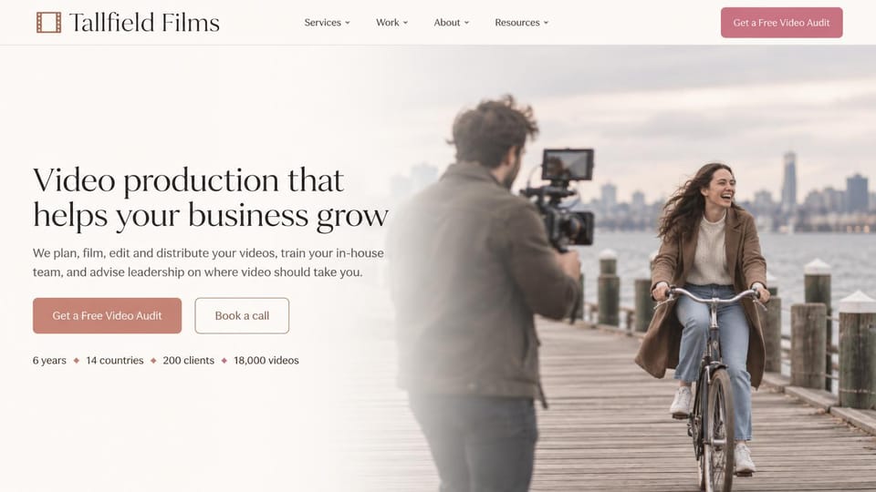 Videographer website design, business video production with a free audit: cream page, dark serif headline, rose audit button and an outlined call button, a filmmaker recording a laughing woman cycling on a waterfront boardwalk