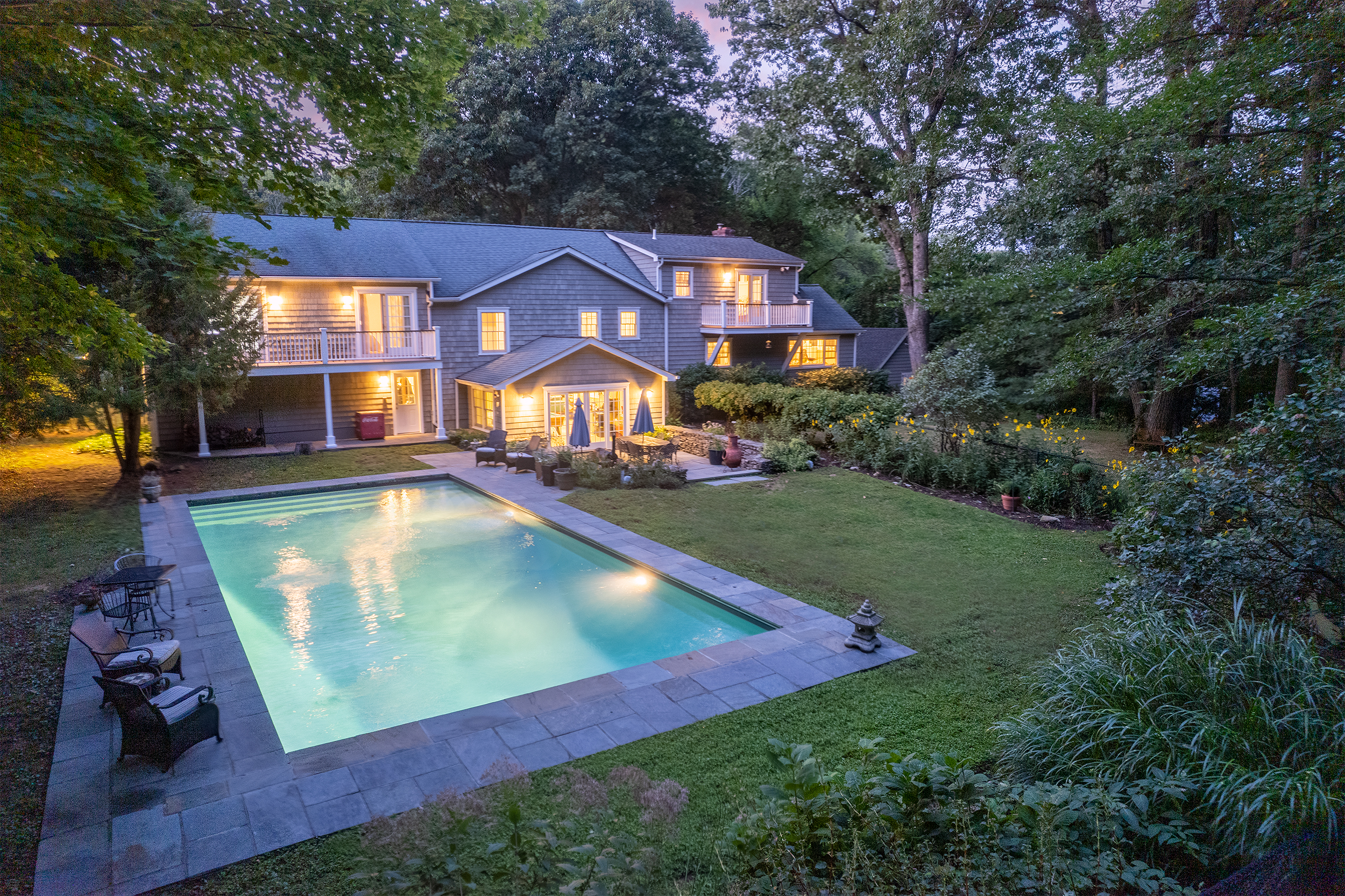 Twilight listing photo of a shingled colonial backyard with a glowing lit pool under a violet dusk sky